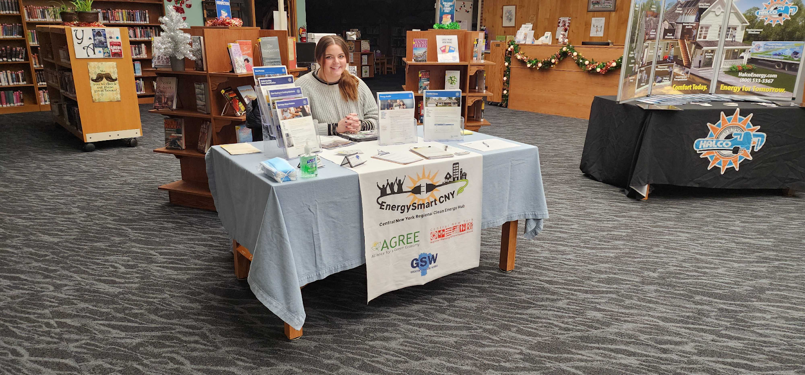 Alt text: Lacey sits smiling at a table covered by a banner showcasing the organizations that make up the Central New York Hub. There are also a number of flyers, brochures, and educational materials on the table. The table is staged inside a library; there are bookshelves in the background.